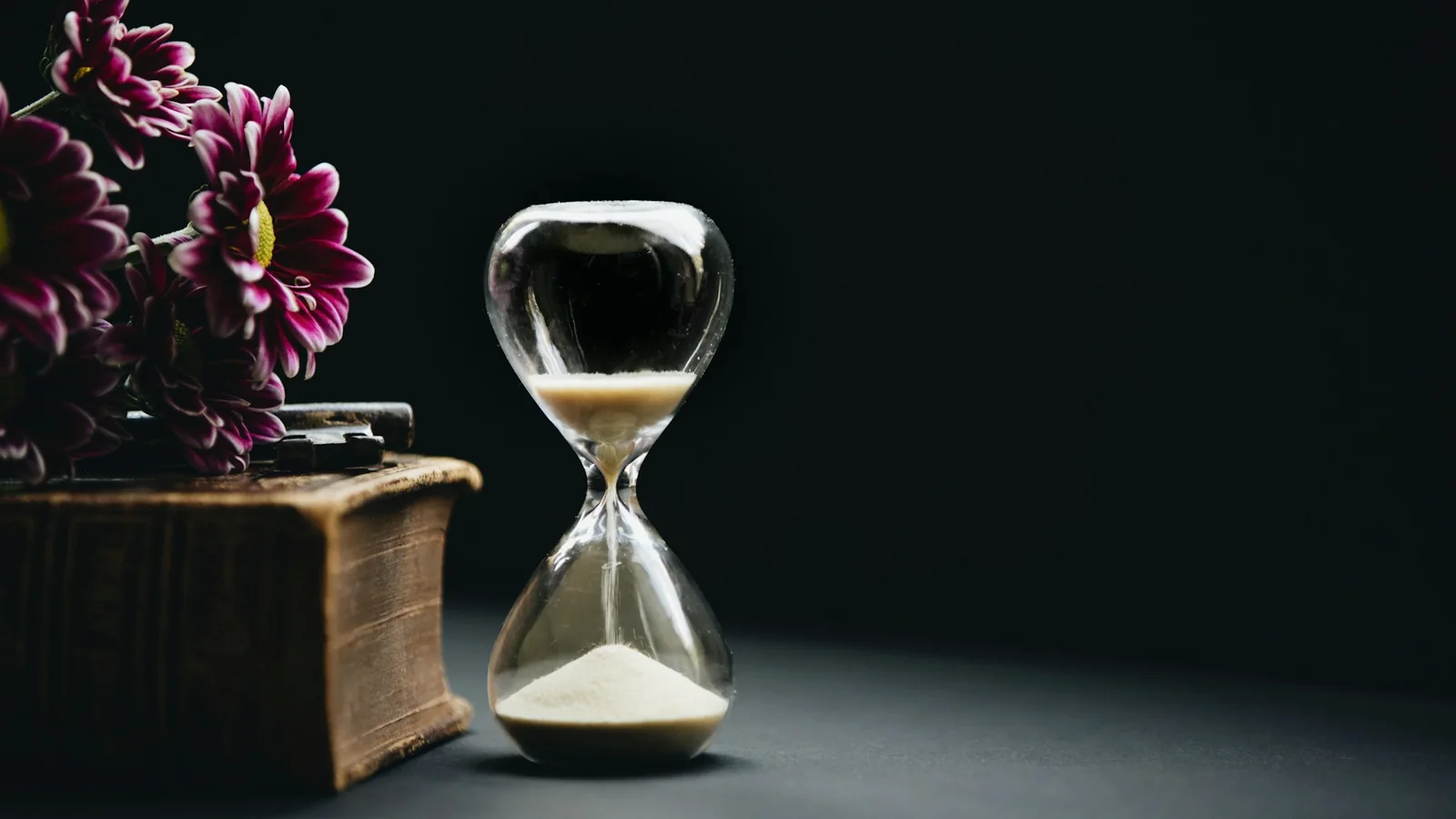 Sablier posé sur un livre ancien, devant des fleurs sombres, fond noir